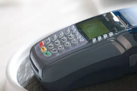 Close-up of a dark gray VeriFone card terminal with a numeric PIN keypad, red, yellow and green function keys and a small green display, resting in a white holder