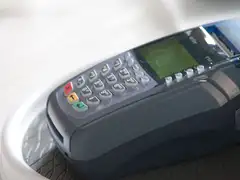 Close-up of a dark gray VeriFone card terminal with a numeric PIN keypad, red, yellow and green function keys and a small green display, resting in a white holder