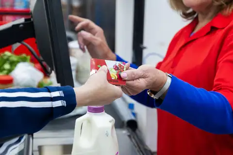 A shopper in a striped jacket hands a red benefits card printed with fruit and vegetables to a cashier in a red shirt at a supermarket checkout, with a gallon of milk on the counter