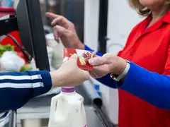A shopper hands a benefits card printed with fruit and vegetables to a cashier at a supermarket checkout, with a gallon of milk on the counter
