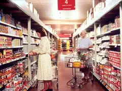 Two women with shopping carts in a supermarket aisle lined with canned goods, under a red hanging sign reading 4, ready to serve, international