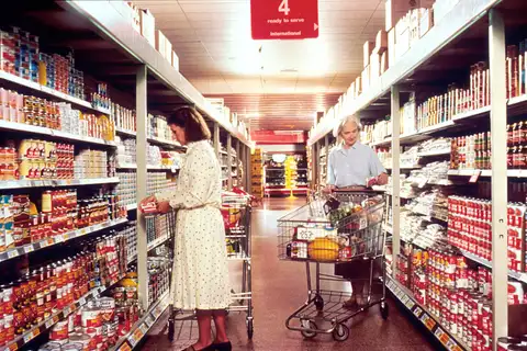 Two women with shopping carts in a supermarket aisle lined with canned goods, under a red hanging sign reading 4, ready to serve, international