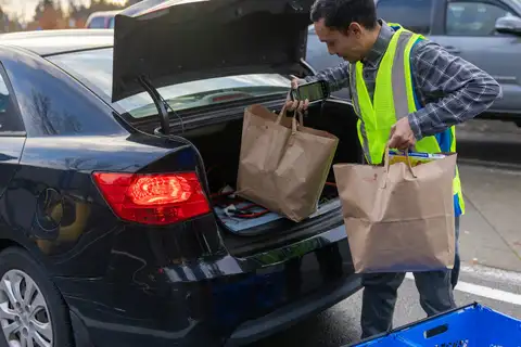 A store worker in a yellow safety vest holds a handheld scanner and lifts paper grocery bags into the open trunk of a black car in a parking lot