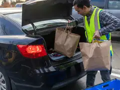 A store worker in a yellow safety vest holds a handheld scanner and lifts paper grocery bags into the open trunk of a black car in a parking lot