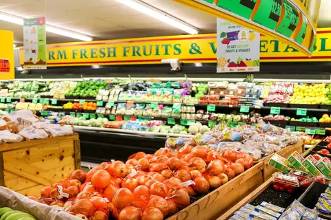 Grocery store produce section with bagged onions in wooden bins, shelves of fruit and vegetables, and hanging signs offering 50% off fruits and veggies