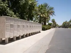 A row of beige cluster mailboxes on posts beside a sidewalk on a sunny street with palm trees and parked cars