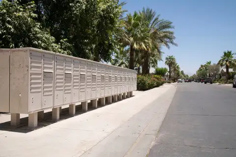 A row of beige cluster mailboxes on posts beside a sidewalk on a sunny street with palm trees and parked cars