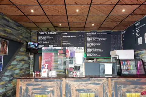Order counter of a small burger shop with chalkboard menus for chicken burgers, hot dogs, sides, burgers, drinks and shakes, and a cooler of canned sodas