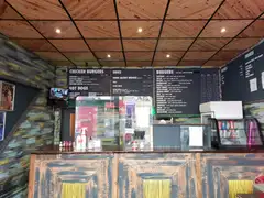 Order counter of a small burger shop with chalkboard menus for chicken burgers, hot dogs, sides, burgers, drinks and shakes, and a cooler of canned sodas
