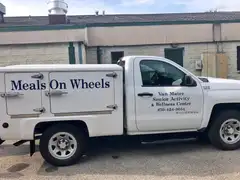 A white pickup truck with an insulated cargo box marked Meals On Wheels, its door lettered Van Matre Senior Activity & Wellness Center, parked beside a low brick building