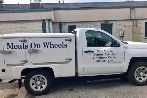 A white pickup truck with an insulated cargo box marked Meals On Wheels, its door lettered Van Matre Senior Activity & Wellness Center, parked beside a low brick building