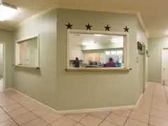 Reception windows with sliding glass panels set into a pale green wall in a clinic lobby with a tiled floor, exit signs and a staff member at a desk behind the glass