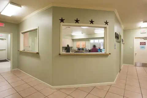Reception windows with sliding glass panels set into a pale green wall in a clinic lobby with a tiled floor, exit signs and a staff member at a desk behind the glass