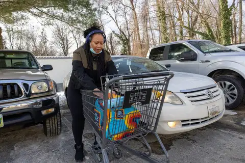 A woman wearing headphones unloads grocery bags from a shopping cart between parked cars in a parking lot with patches of snow