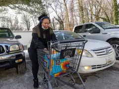A woman wearing headphones unloads grocery bags from a shopping cart between parked cars in a parking lot with patches of snow