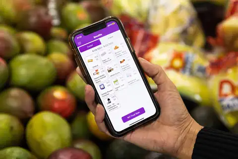 A hand holds a smartphone showing a purple WIC benefits app screen that lists current food benefits for 4/24/2023 to 5/23/2023, such as cheese, eggs and whole grains, in front of a blurred display of mangoes