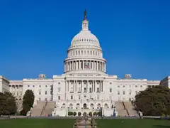 The west front of the United States Capitol in Washington, D.C., with its white dome and two long wings under a clear blue sky, seen across a lawn with trees