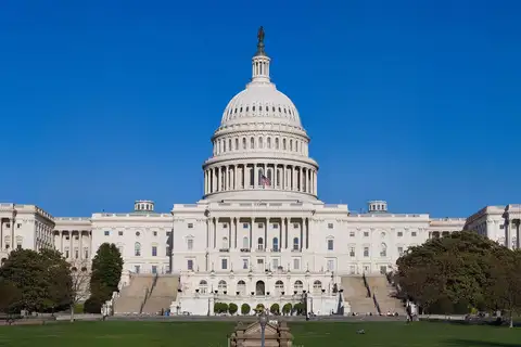 The west front of the United States Capitol in Washington, D.C., with its white dome and two long wings under a clear blue sky, seen across a lawn with trees